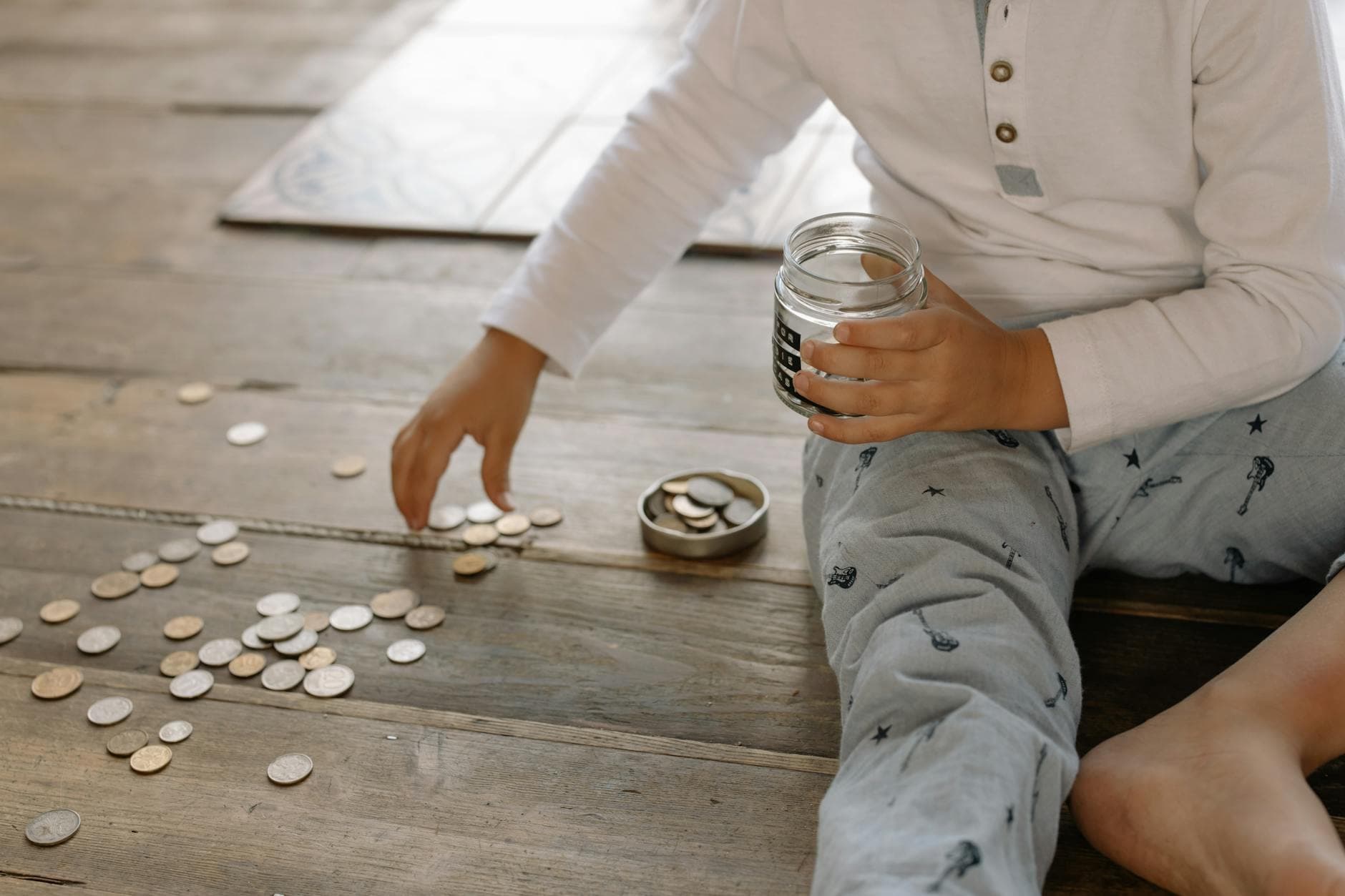 Why Mental Accounting Makes You Spend More Money — A young child collects coins into a jar on a wooden floor, symbolizing savings.